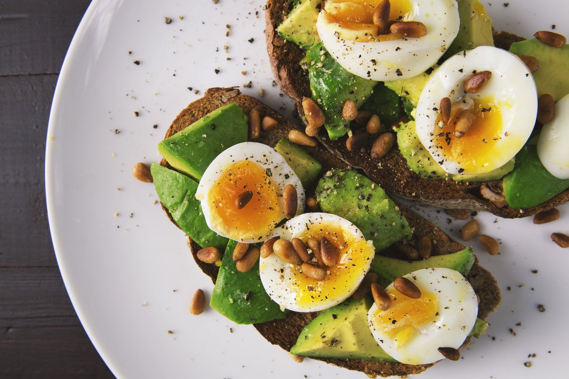 Avocado toast with soft-boiled eggs, sprinkled with seeds and pepper, on a white plate.